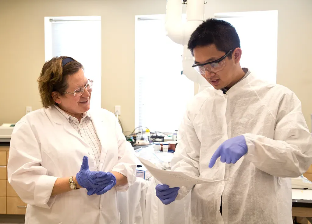 A student in a white lab coat talks to a faculty member